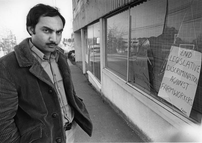 [Portrait of Raj Chouhan stands in front of vandalized storefront office window]
