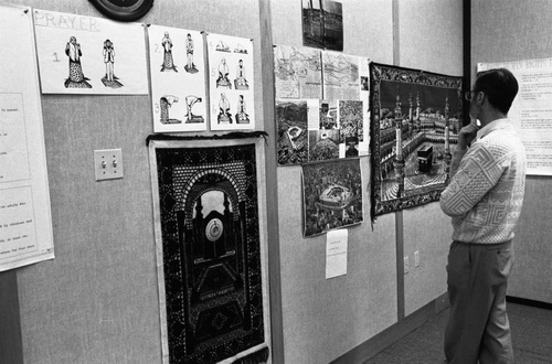 A man in a sweater and glasses stands in front of a wall adorned with various posters and prayer rugs, including diagrams of prayer postures and images of Mecca.