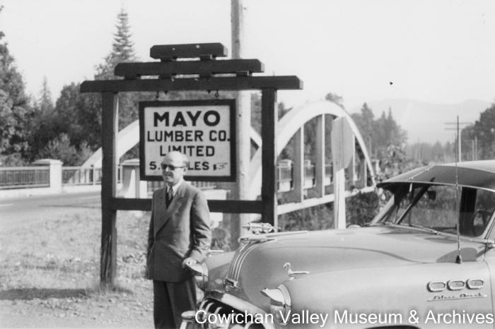 [Carl Friegon posing with a car in front of a bridge and sign for May Lumber Co.]