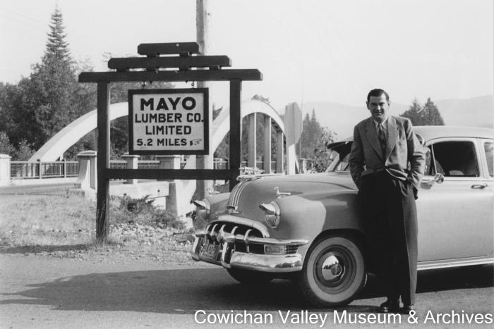 [Henry McHemer posing with a car in front of a bridge and sign for May Lumber Co.]
