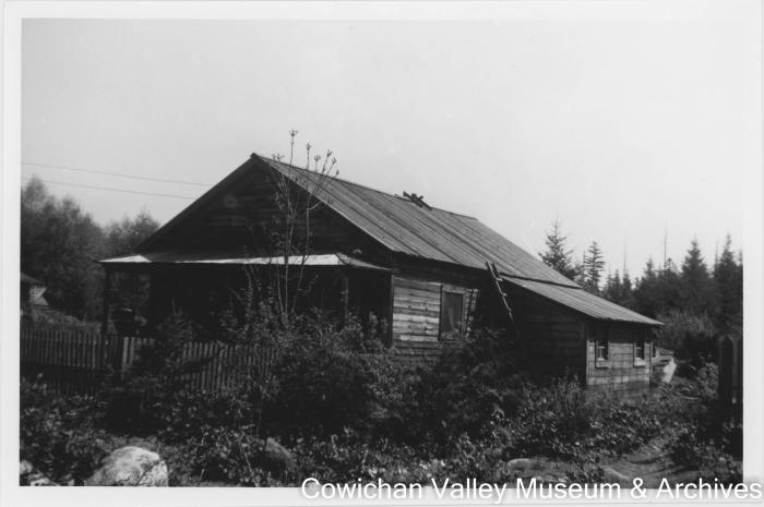 [A wooden house surrounded by bushes and trees in the background]