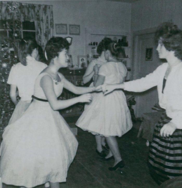 [Photo of a group of women dancing in pairs inside a house]