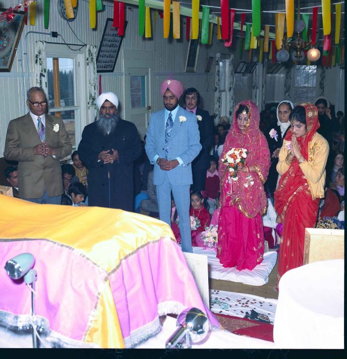[Group photo of Braich and unidentified  bride with Head Granthi standing and doing Ardaas in front of the sikh Sri Guru Granth Sahib]
