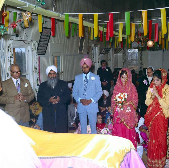 [Group photo of Braich and unidentified  bride with Head Granthi standing and doing Ardaas in front of the sikh Sri Guru Granth Sahib]