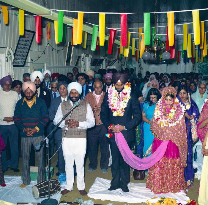 [Group photo of Balbir Khangura and Baljit Gill with Head Granthi standing and doing Ardaas in front of the sikh Sri Guru Granth Sahib]