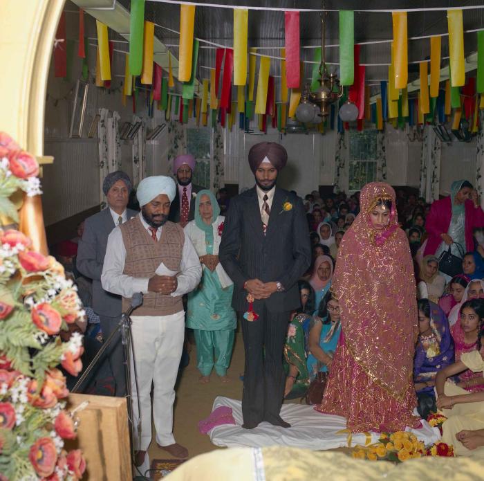 [Group photo of Balbir Khangura and Baljit Gill with Head Granthi standing and doing Ardaas in front of the sikh Sri Guru Granth Sahib]