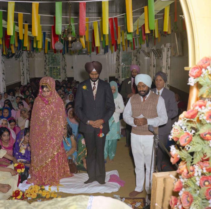 [Group photo of Balbir Khangura and Baljit Gill with Head Granthi standing and doing Ardaas in front of the sikh Sri Guru Granth Sahib]