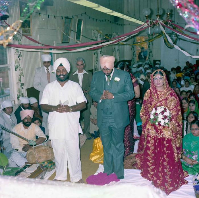 [Group photo of Inderjit Cheema and unidentified bride with Head Granthi standing and doing Ardās in front of the Sri Guru Granth Sahib]