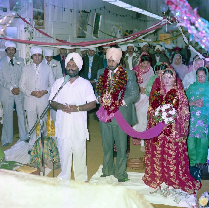 [Group photo of Inderjit Cheema and unidentified bride with Head Granthi standing and doing Ardās in front of the Sri Guru Granth Sahib]