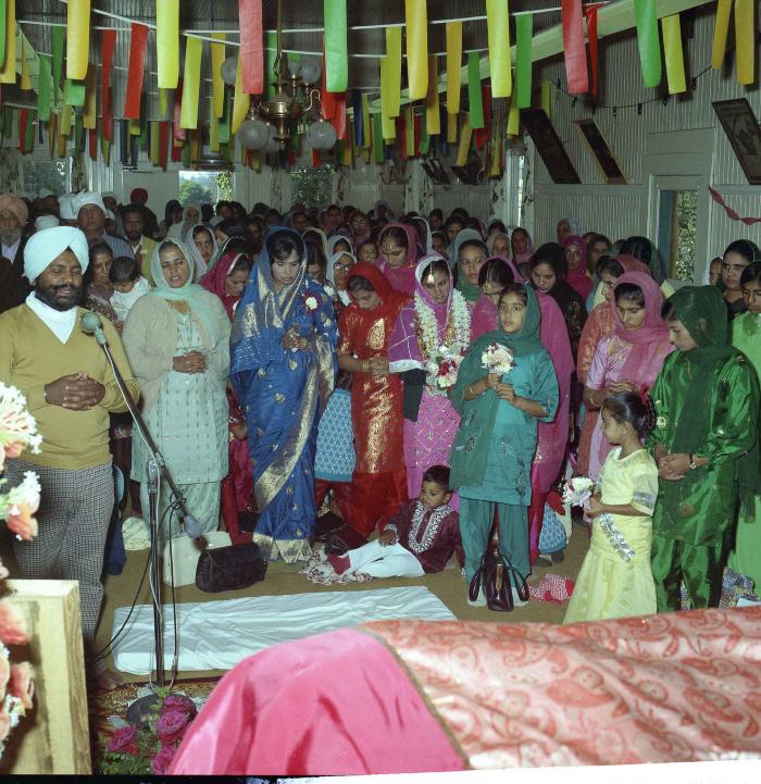 [Group photo of unidentified bride and wedding guests with Head Granthi standing and doing Ardās in front of the Sri Guru Granth Sahib]