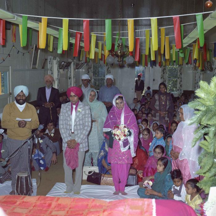 [Group photo of unidentified bride and wedding guests with Head Granthi standing and doing Ardās in front of the Sri Guru Granth Sahib]